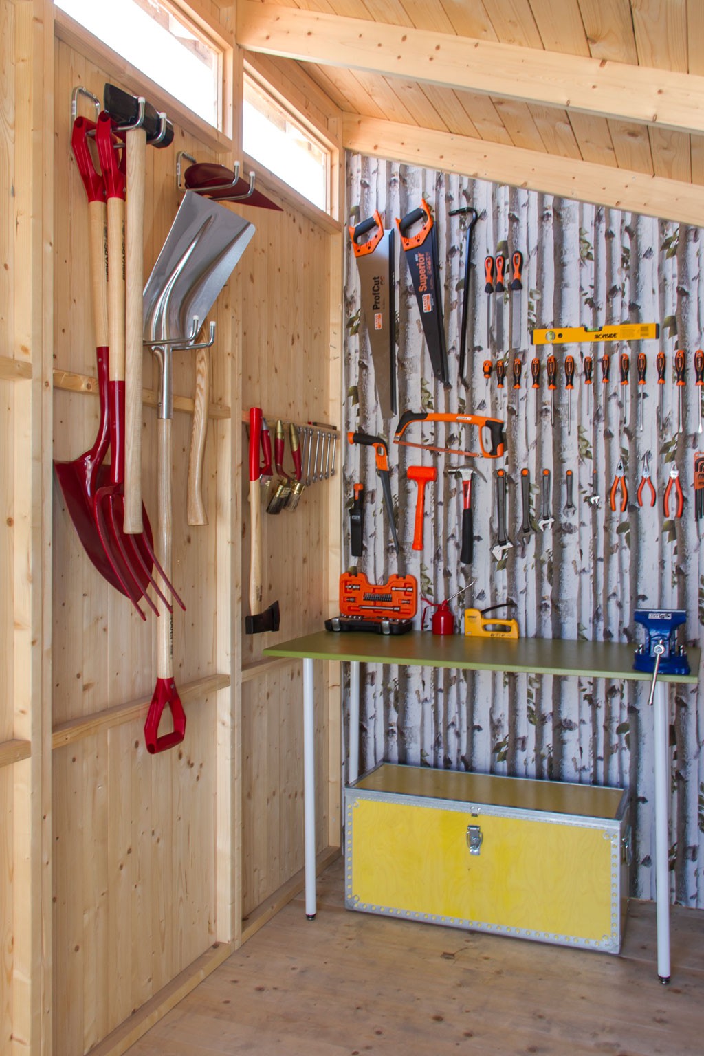 Interiør i lyst treverk med redskaper og sager hengende, birkekledning, grønt bord og gul kasse.