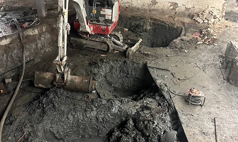 Indre byggeplass med rød-hvitt minigraver, gjørmete gulv og dype firkantede hull.