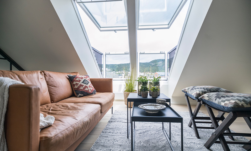 Lyst loftrom med brun skinnsofa, svart bord med grønne planter, grått teppe og byutsikt