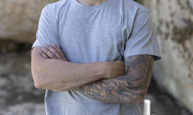 Nærbilde av kryssede armer i grå t-skjorte, høyre arm med svartgrå fullarmstatovering.