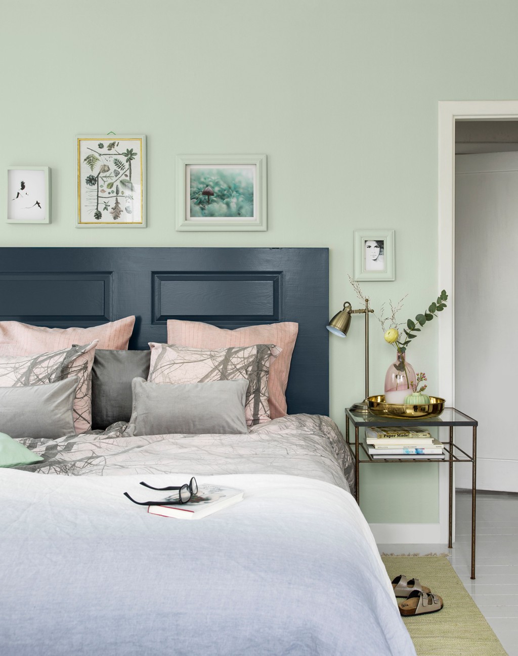 Pastellgrønt soverom med mørkeblå sengegavl, rosa/grå puter, glassbord med messinglampe.