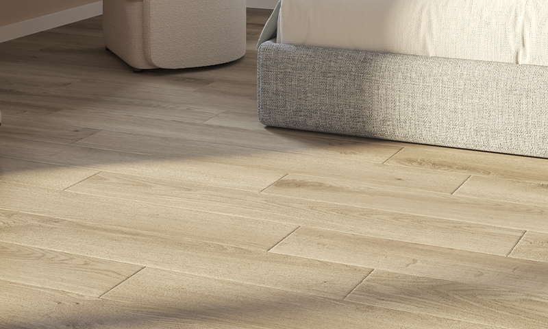 Lyst eikeparkettgulv under grå sengebunn og kremfarget puff badet i dagslys