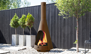 Brun metallpeis med ild på grus i moderne utehage foran svart plankegjerde.