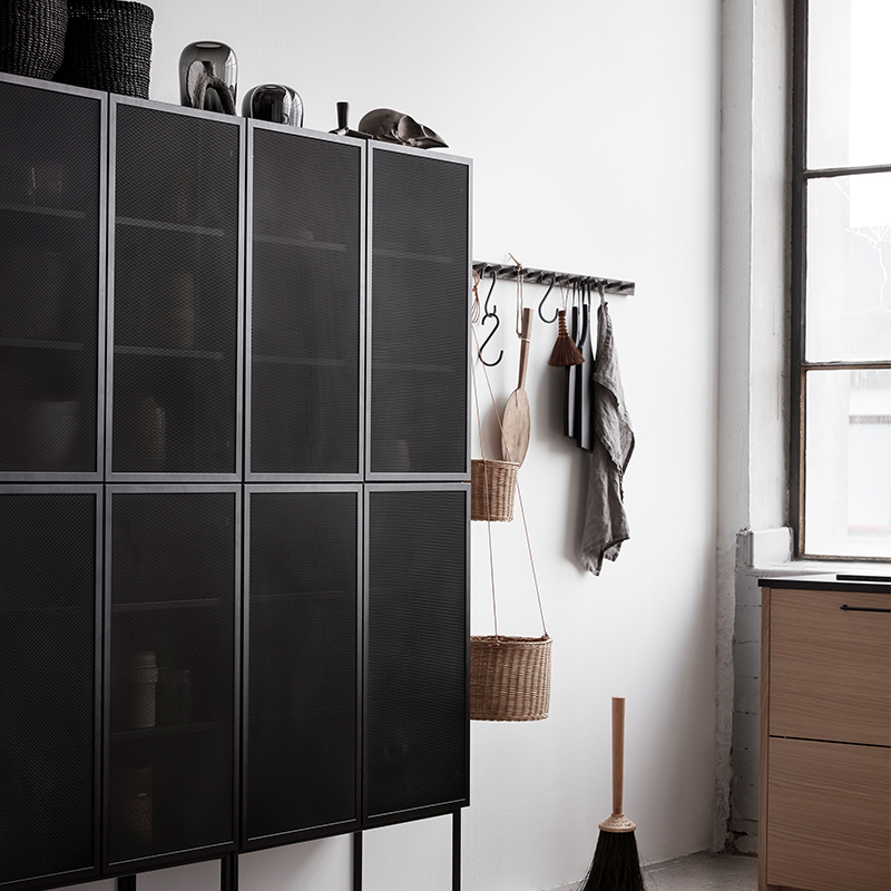 Svart metallnetting-skap mot hvit vegg, hengende kurver, tresleiv, kost og vindu.