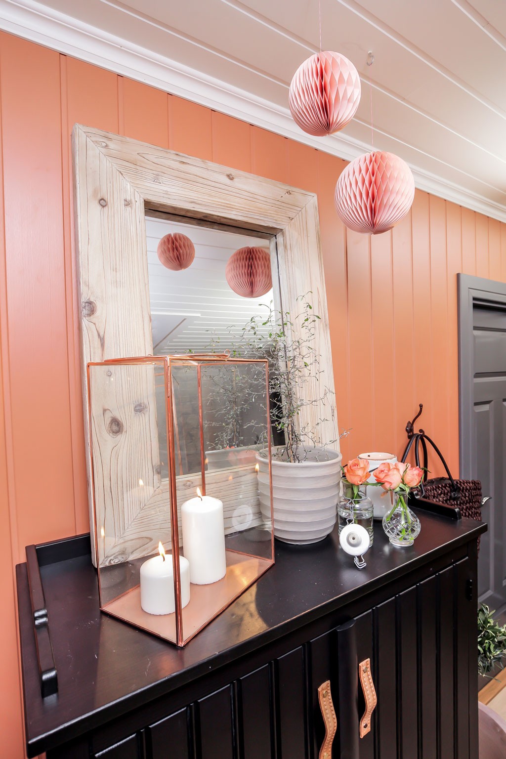 Svart konsollbord foran korallfarget vegg med tre papirkuler, speil, tente lys og roser.