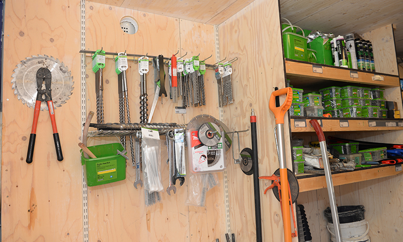 Trevegg i jernvarebutikk med rød boltesaks, sagblad, bor og grønne oppbevaringsbokser.