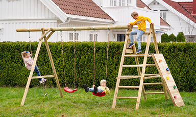 Tre barn leker på lyst treklatrestativ på grønn plen med hvite hus og rødt tak i bakgrunnen