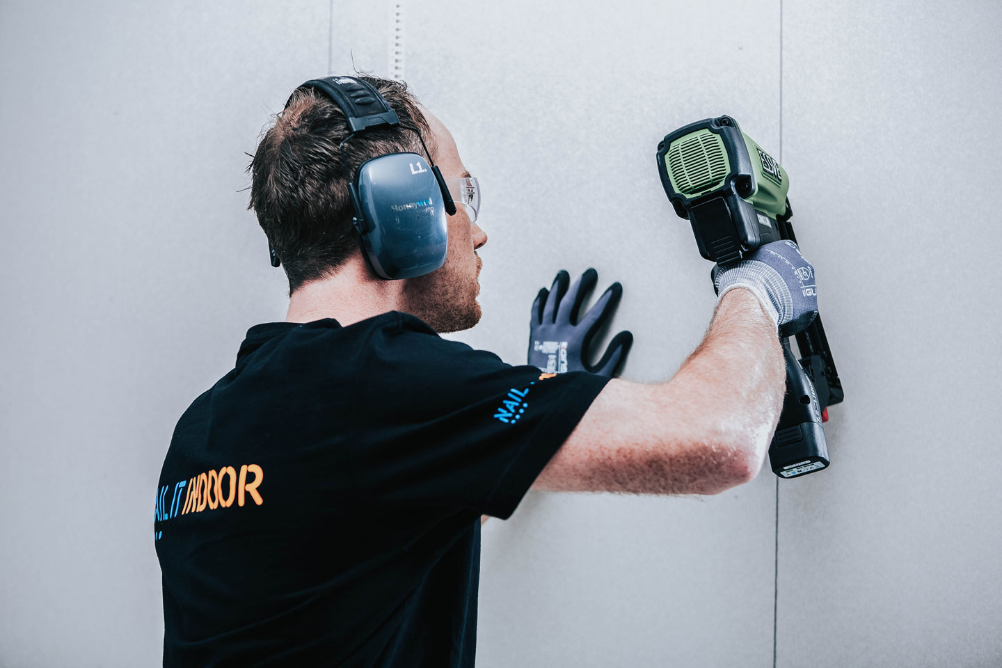 Håndverker monterer gipsplate med grønn spikerpistol iført hørselsvern og hansker.