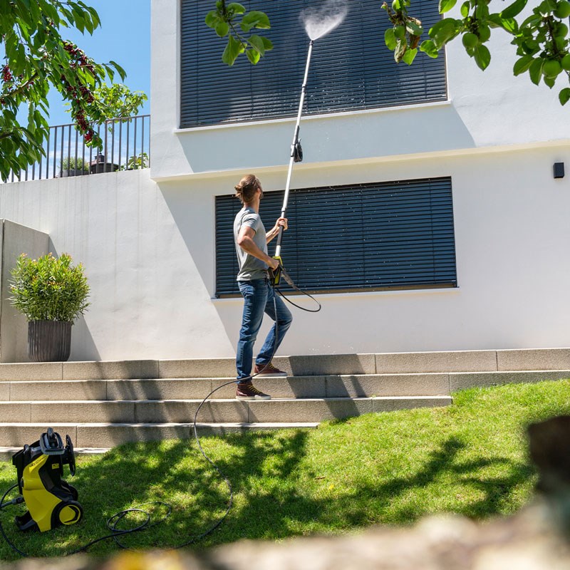 Mann med gul høytrykksvasker rengjør øvre vindusskodder på hvit fasade ved grønt gress.