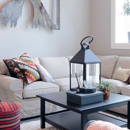 Lyst stuehjørne med beige sofa, flerfarget puter, svart bord med lykt og planter.