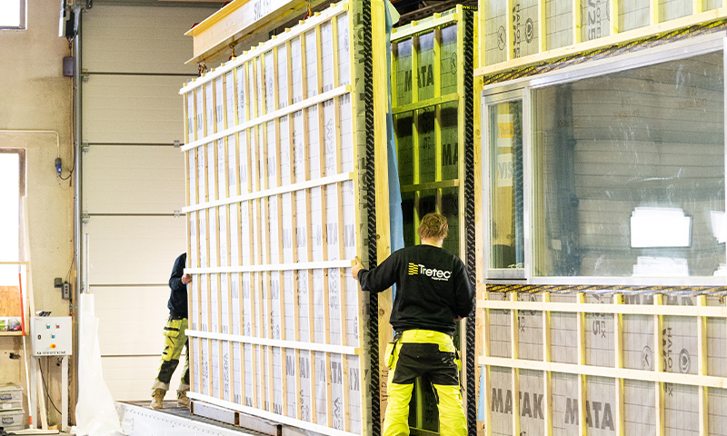 To håndverkere i gule vernebukser manøvrerer en stor hvitgul isolert vegg i lyst verksted.