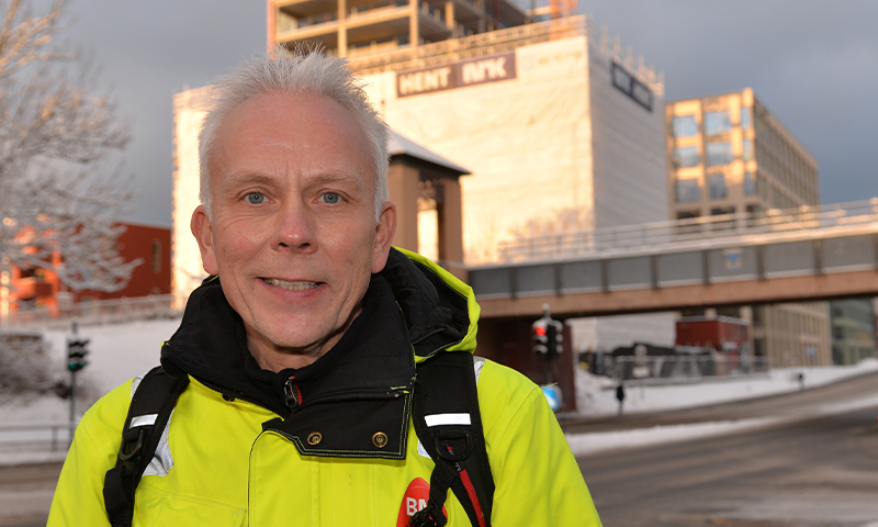 Mann i gul arbeidsjakke med ryggsekk står i snødekt bygate foran kontorbygg og gangbro.