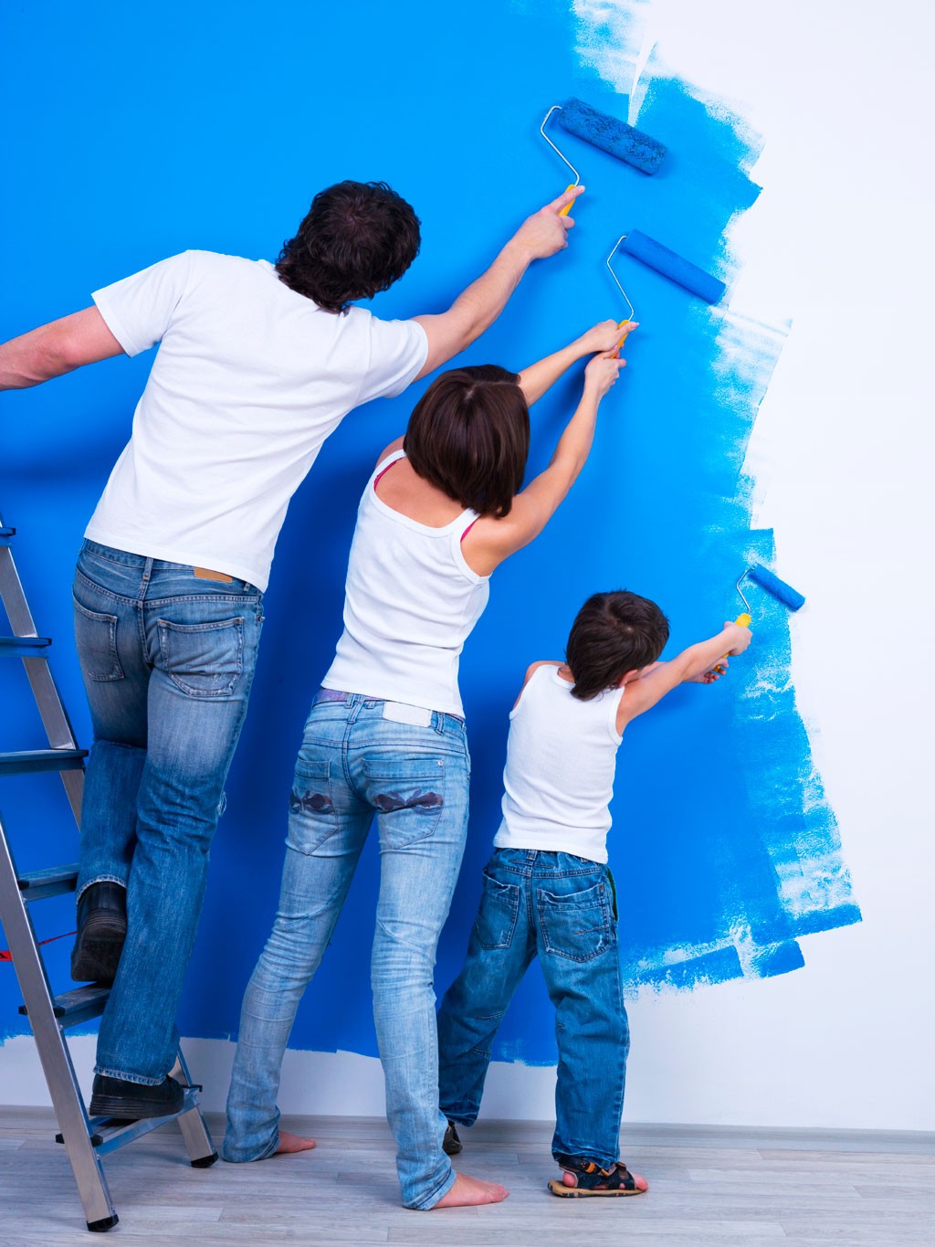 Far på stige, mor og barn maler vegg blå med maleruller, alle i hvite topper og blå jeans.