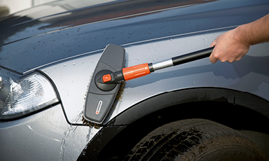 Hånd vasker sølvgrå bil ved hjulbuen med svart-oransje børste og vannperler.