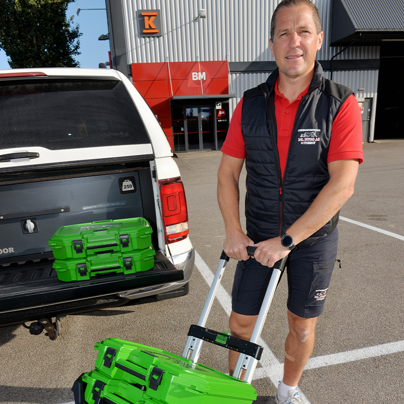 Håndverker med rød skjorte og grønne verktøykasser ved hvit pickup utenfor BM-bygning