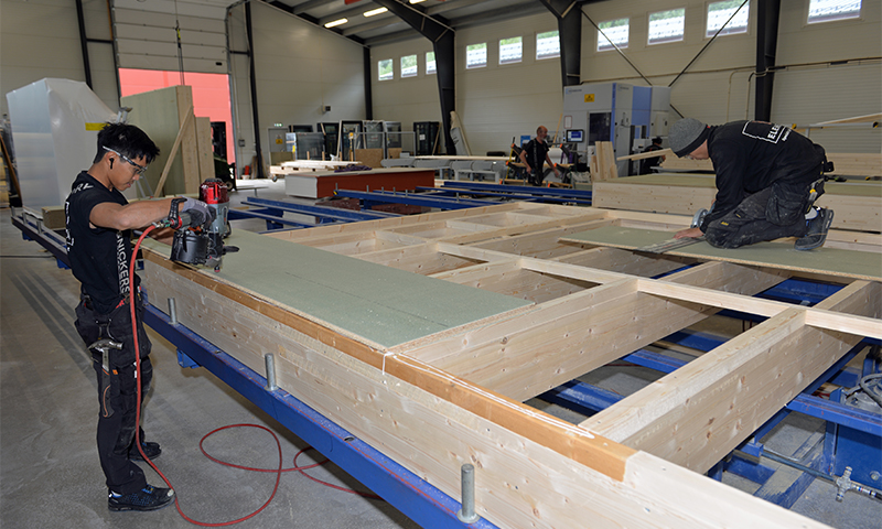 To snekkere bygger store lyse trekonstruksjoner på blå monteringsramme inne i verksted 