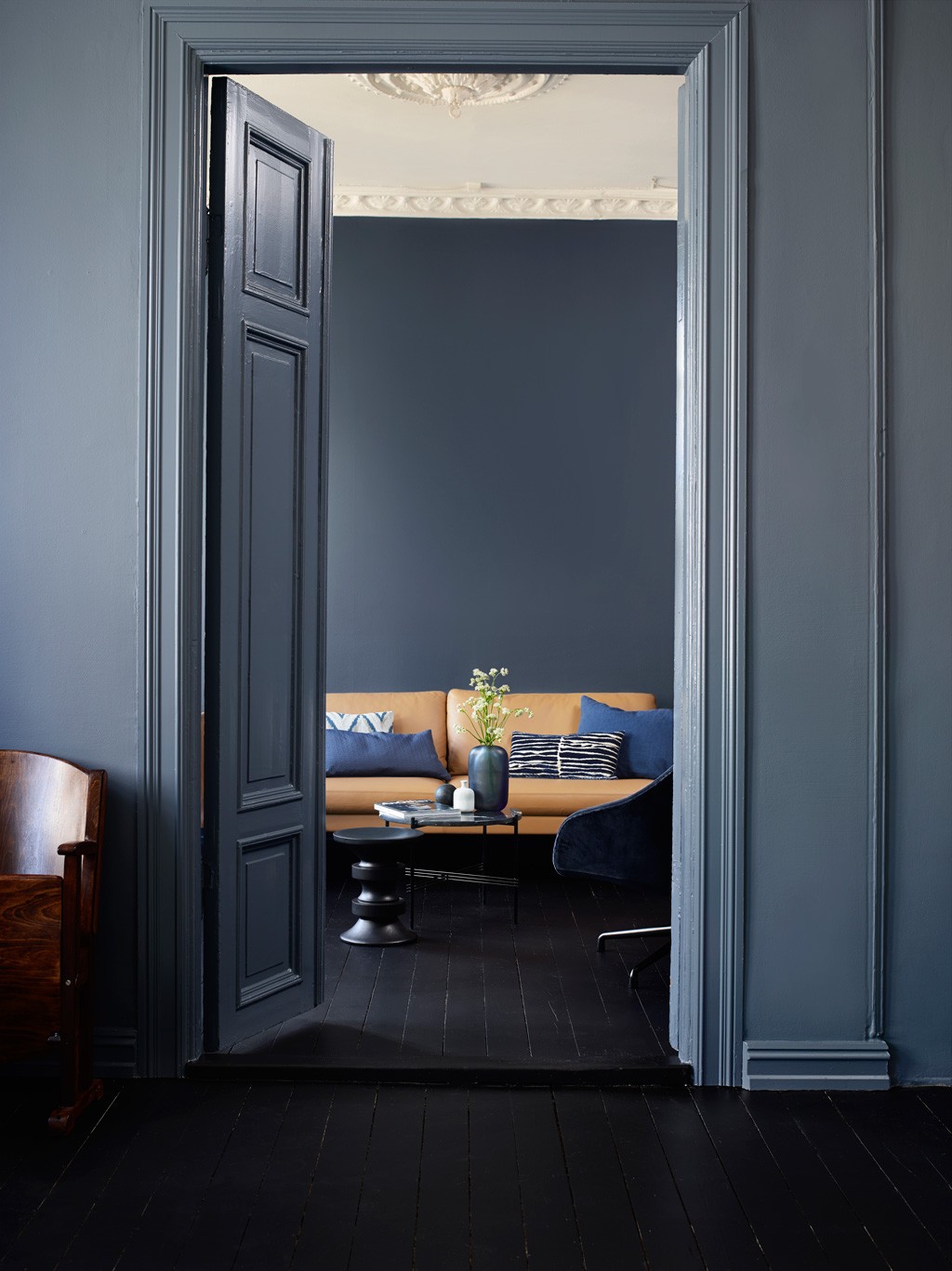 Åpen dør til stue med mørkeblå vegger, svart gulv, brun sofa med blå puter og vase med blomster.