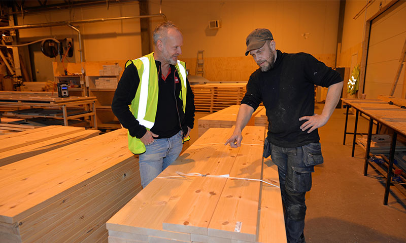 To menn i treverksted under oransje lys, én i gul vest, peker på stabler av lyst tre.