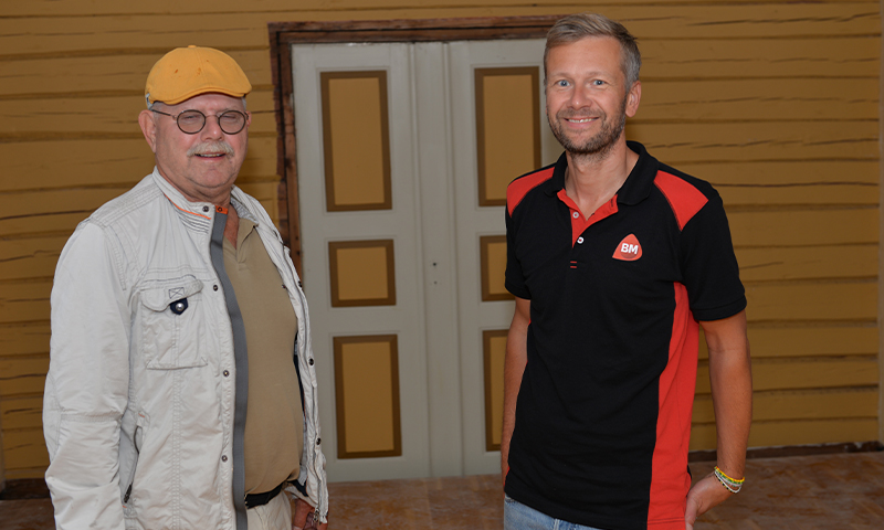 To menn (sort-rød skjorte og lys jakke) foran okergul tømmervegg med hvit-gul dobbeltdør.