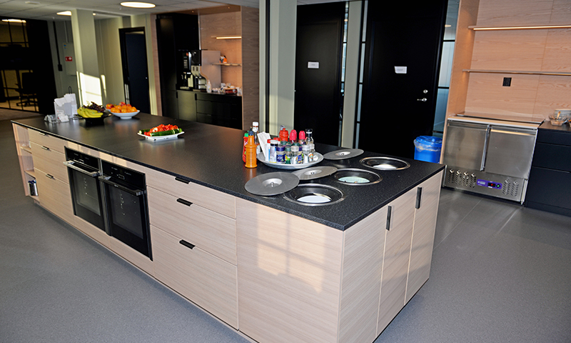 Moderne felleskjøkkenøy i lyst tre med svart benkeplate, fruktfat, kaffemaskin og resirkulering.