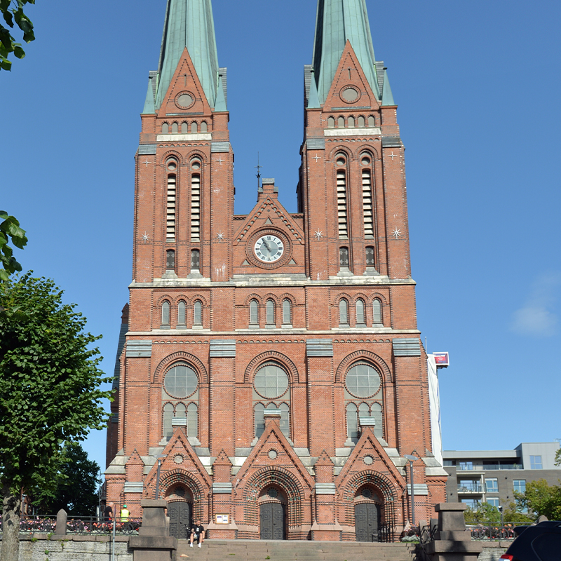 Rød teglsteinskirke med to grønne spir, rundt klokkevindu og klar blå himmel.