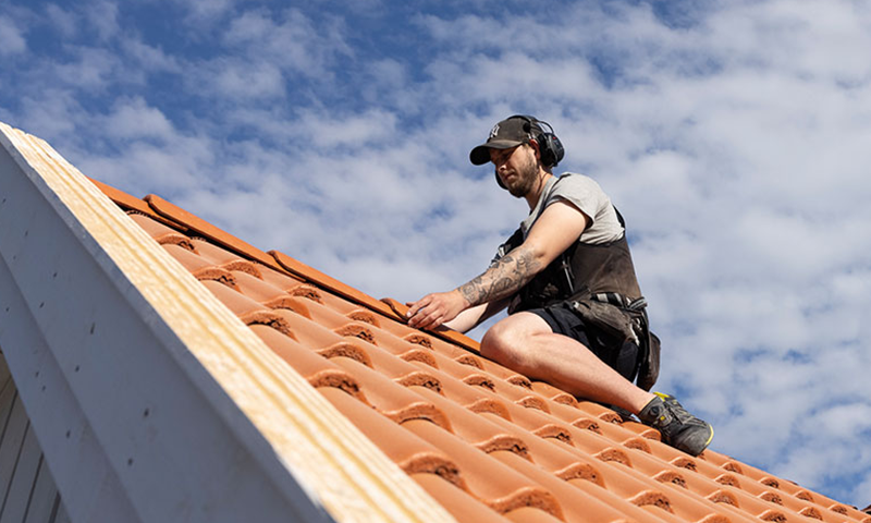 Taktekker i verneutstyr legger rød terrakottatakstein på hvitt hus under blå, skyet himmel