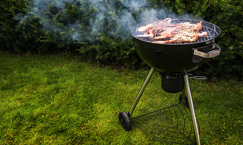 Svart kullgrill med rykende hummerhaler og reker på grønn gressplen foran hekk.