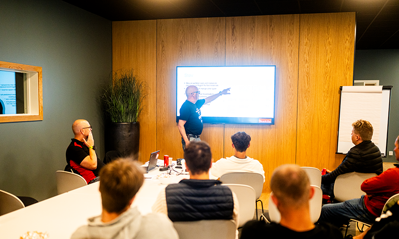 Konferanserom med trepanel, hvite møbler, deltakere rundt bord mens noen presenterer på skjerm