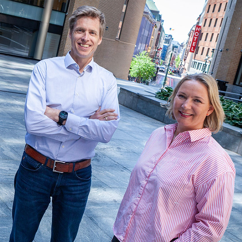 To personer smiler i bymiljø med bygninger i bakgrunnen, en i hvit skjorte og en i rosa stripet skjorte.