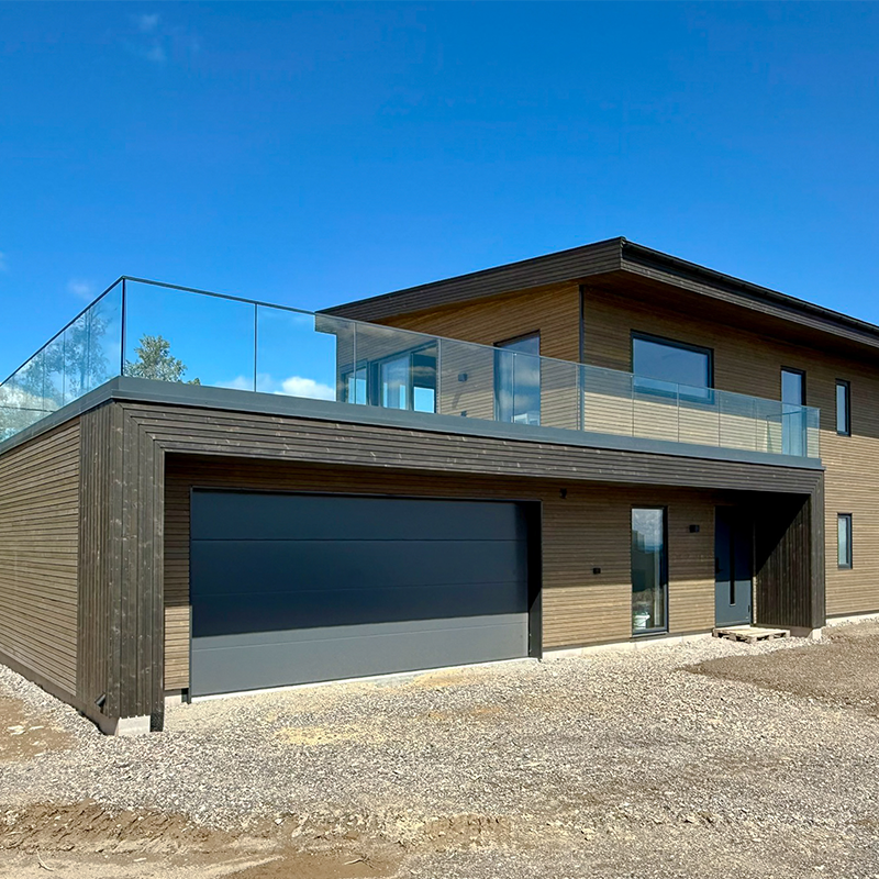 Moderne trehus med garasje, glassbalkong og trepanel fasade mot klar blå himmel, omgitt av gruslagt tomt.