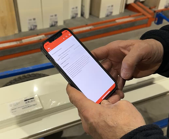 To hender holder smarttelefon med oransje-hvitt app-skjerm foran hyller med byggeplater