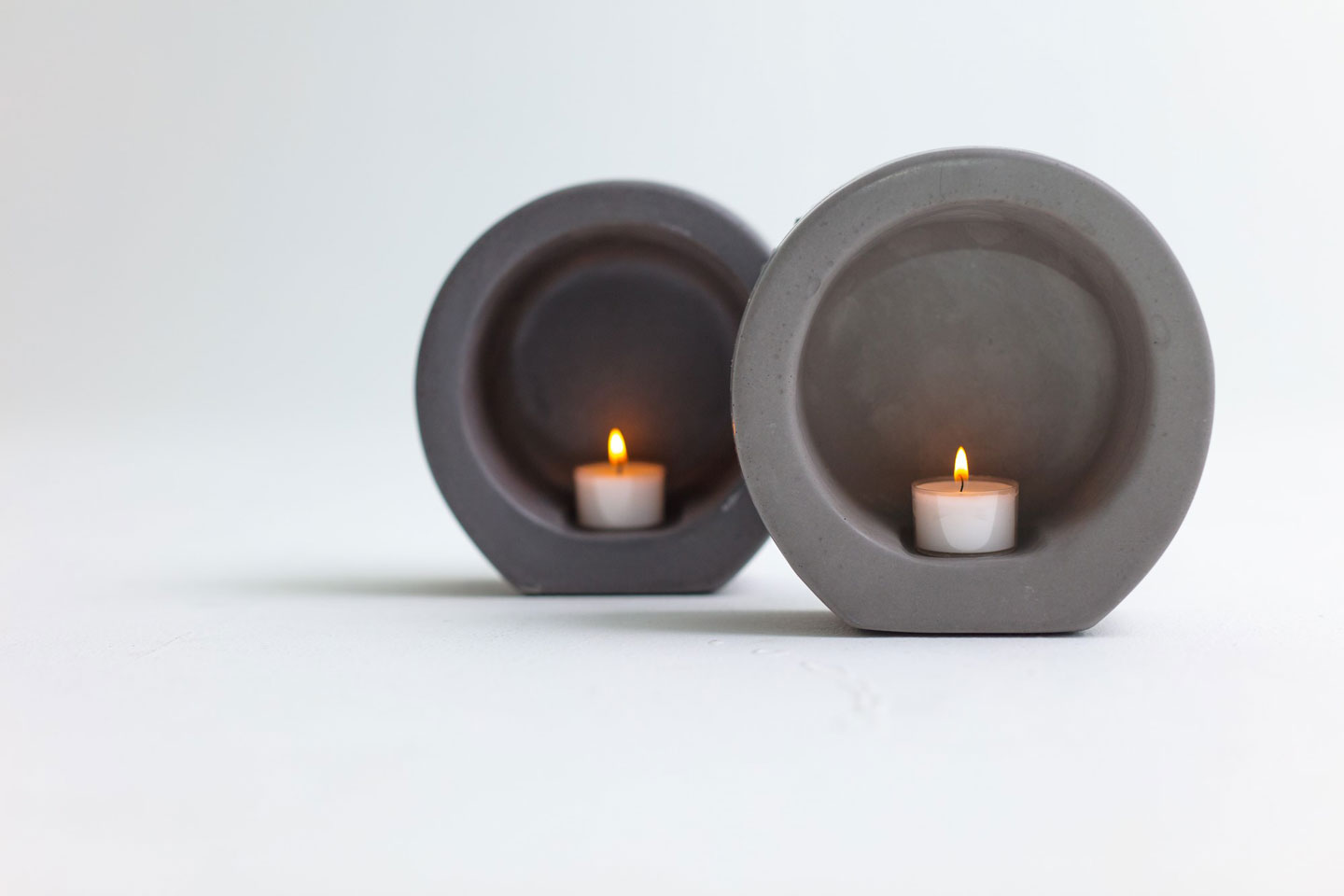 To sirkulære, grå betonglysestaker med brennende telys mot hvit bakgrunn.