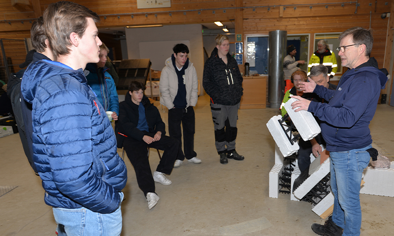 Ungdommer i jakker lytter til instruktør som viser hvite byggeplater i treinteriør.