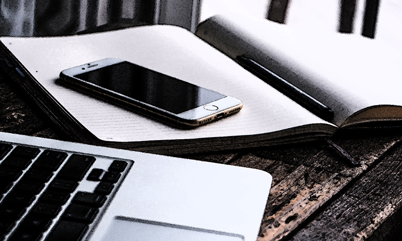 Smarttelefon på åpen notatbok med penn, laptop på rustikt trebord.