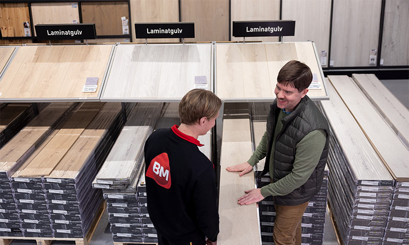 To menn i byggevarebutikk undersøker lyst laminatgulv under skilt “Laminatgulv”