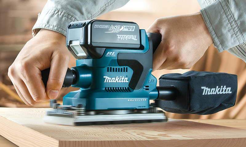 Blå Makita excentersliper i bruk på treplate, to hender holder verktøy og støvpose synlig.