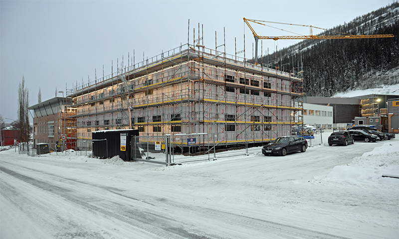 Byggeplass i vinter: nytt trebygg i stillas, gul kran, grå himmel, snø og biler.