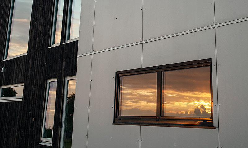 Moderne husfasade i svart tre og grå plater, vindu reflekterer oransje solnedgang.