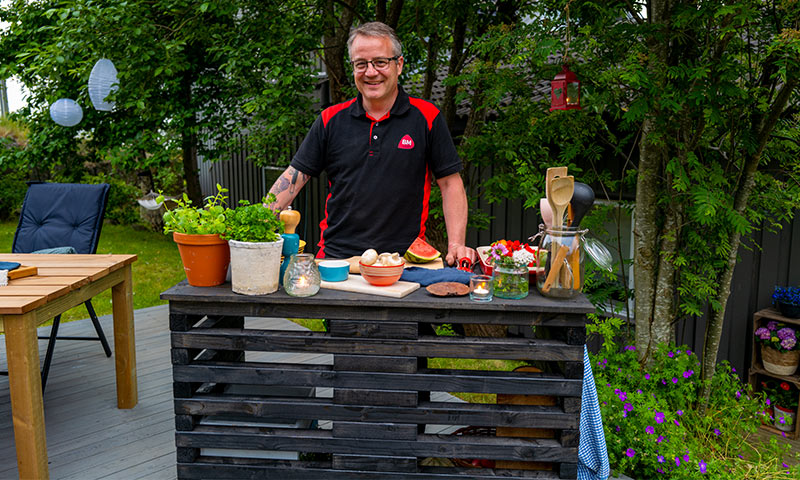 Person i svart-rød skjorte ved svart pallerbar med urter, frukt og redskaper i grønn hage.