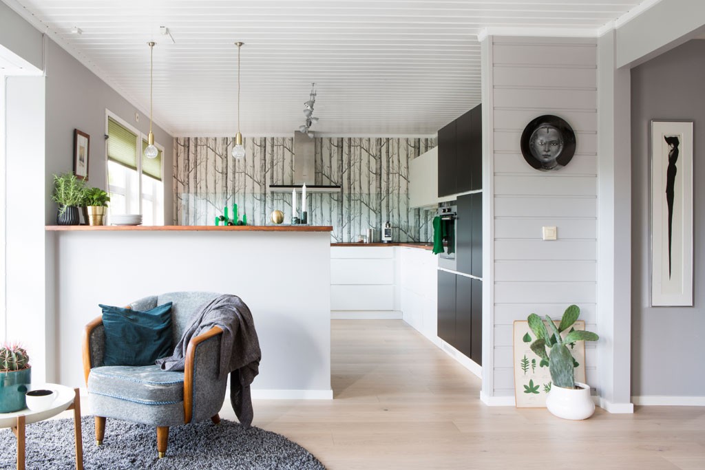 Lyst åpent kjøkken og stue med grå lenestol, hvite skap, bjørkekvist-tapet og grønne planter