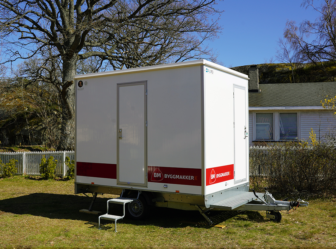 Hvit mobil brakke med rødt Byggmakker-skilt på gressplen, hvitt hus, trær og blå himmel