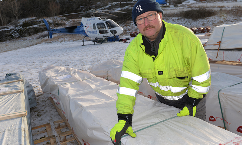 Gult kledd arbeider løfter innpakket trelast på snødekt byggeplass med blått helikopter