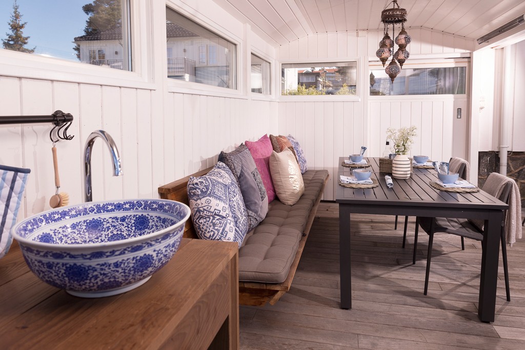 Lyst innglasset uterom med blå-hvitt mønstret vask, benk med fargerike puter og svart spisebord