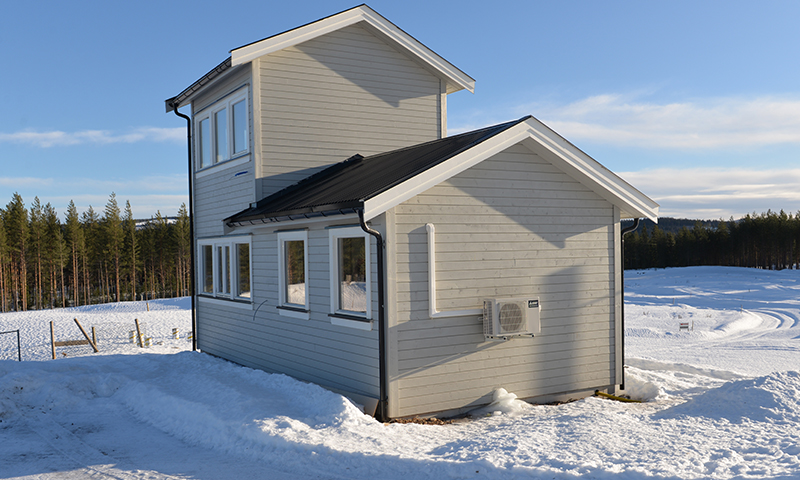 Grått to-etasjes hus med svart tak og hvit varmepumpe, snødekt landskap og furuskog