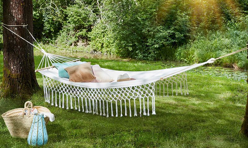 Hvit makramé-hengekøye med pastellputer mellom trær ved grønn hage og dam, kurv på gresset.