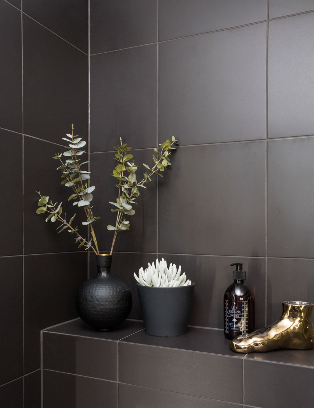 Mørk flishylle med svart vase med eukalyptus, grå potte hvit sukkulent, brun såpeflaske og gullfot