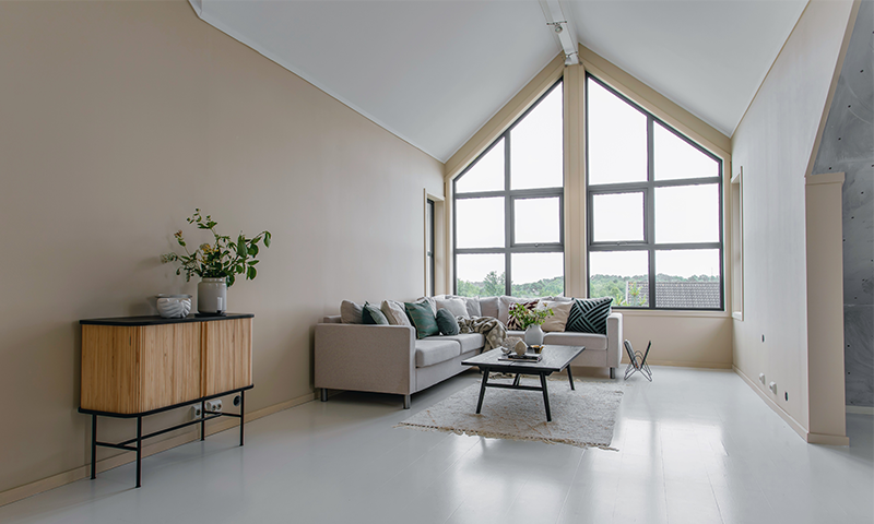 Lys minimalistisk stue med hvitt gulv, beige vegger, hjørnesofa, treskap og store vinduer.