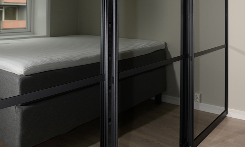 Soverom med mørk grå seng og hvit madrass bak speilglass-skyvedører i sort ramme.