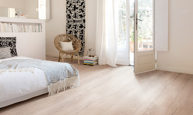 Lyst soverom med hvit seng, grått pledd, rottingstol, bokstabel, sort-hvitt tapet og åpen dør.