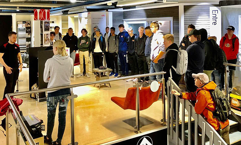 Ungdommer lytter til presentasjon i lyst møbelshowrom med røde stoler og metallrekkverk.